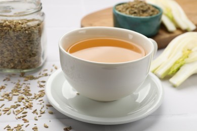 Photo of Fennel tea in cup, seeds and fresh vegetable on white tiled table, closeup