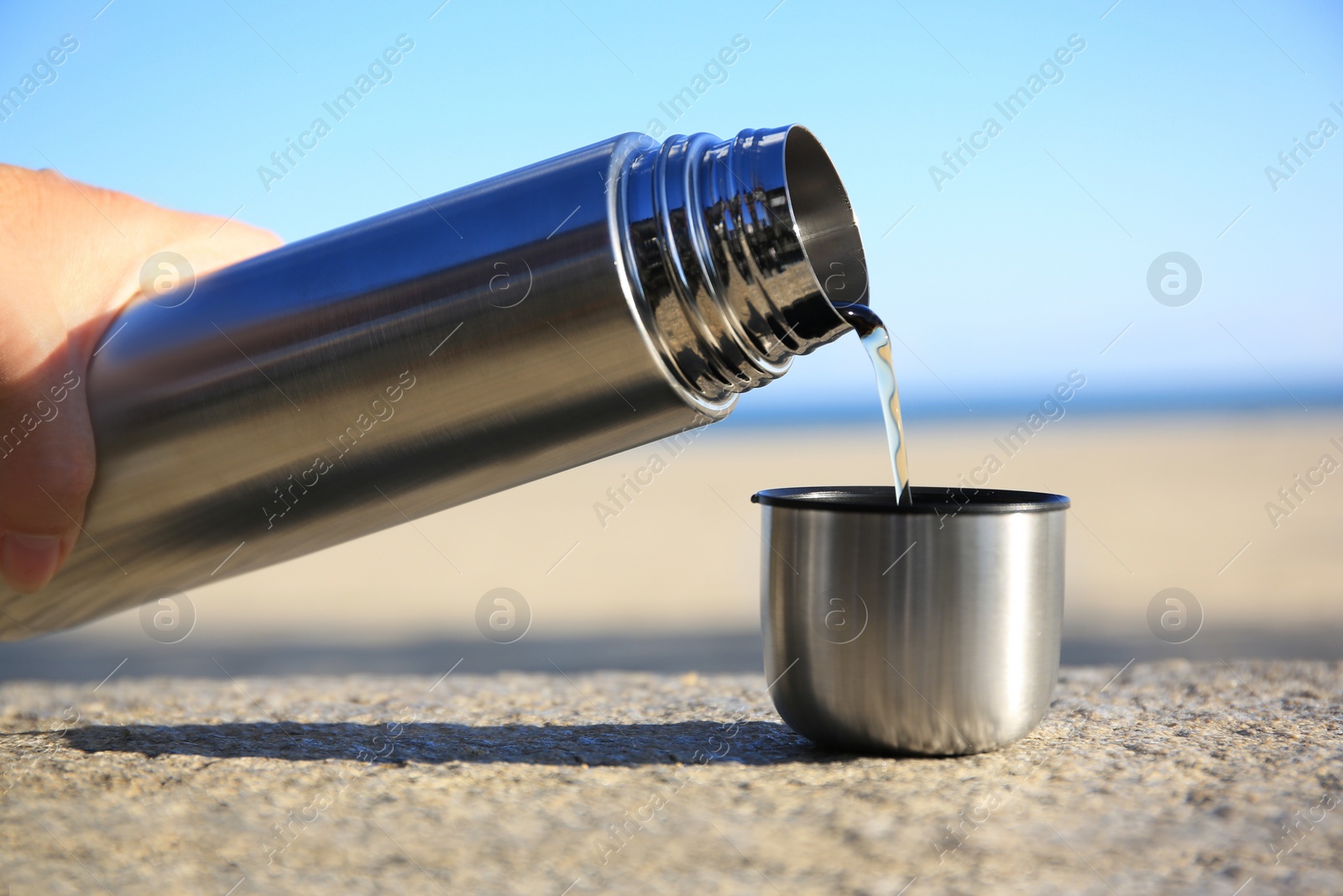 Photo of Woman pouring hot drink from metallic thermos into cap on stone surface outdoors, closeup
