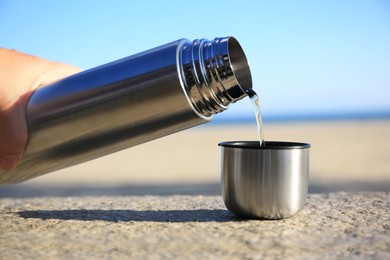 Woman pouring hot drink from metallic thermos into cap on stone surface outdoors, closeup