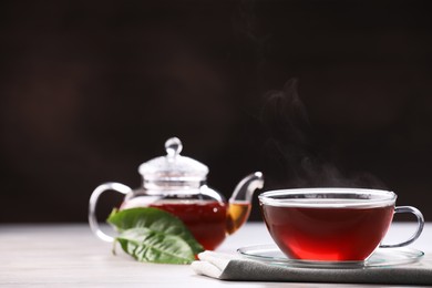 Photo of Aromatic hot tea in glass cup, teapot and leaves on white wooden table against black background. Space for text