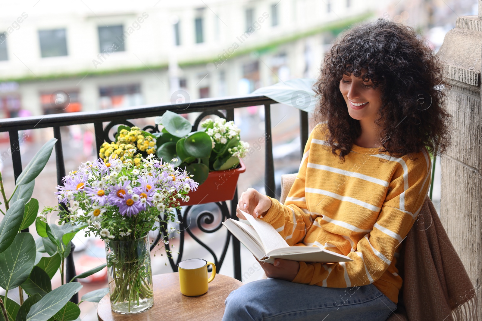 Photo of Young woman reading book at table with beautiful flowers on balcony