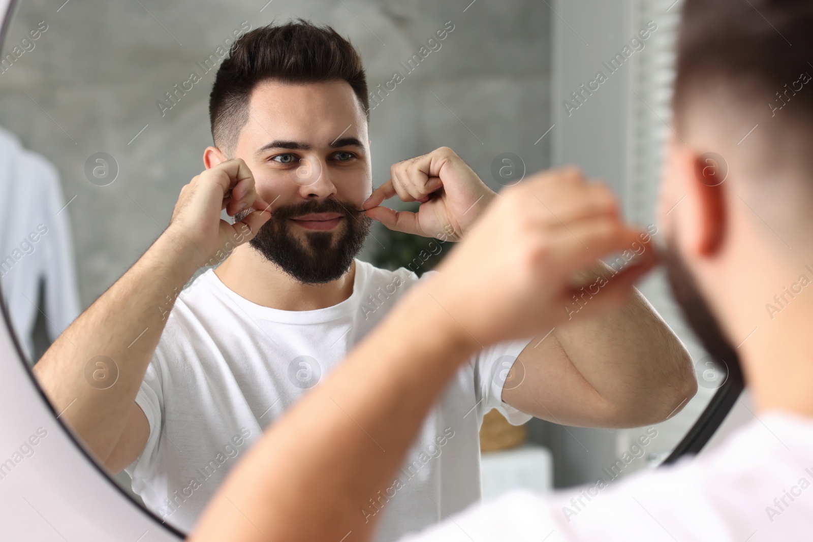Photo of Handsome young man touching mustache near mirror in bathroom