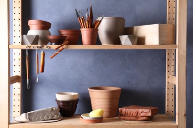 Photo of Set of different crafting tools and clay dishes on wooden rack in workshop