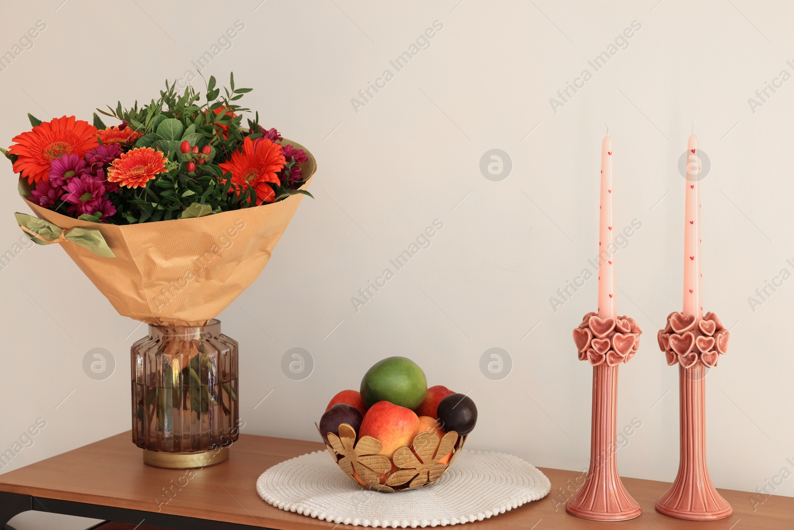 Photo of Bouquet of flowers, bowl with fresh fruits and candles on wooden table near white wall