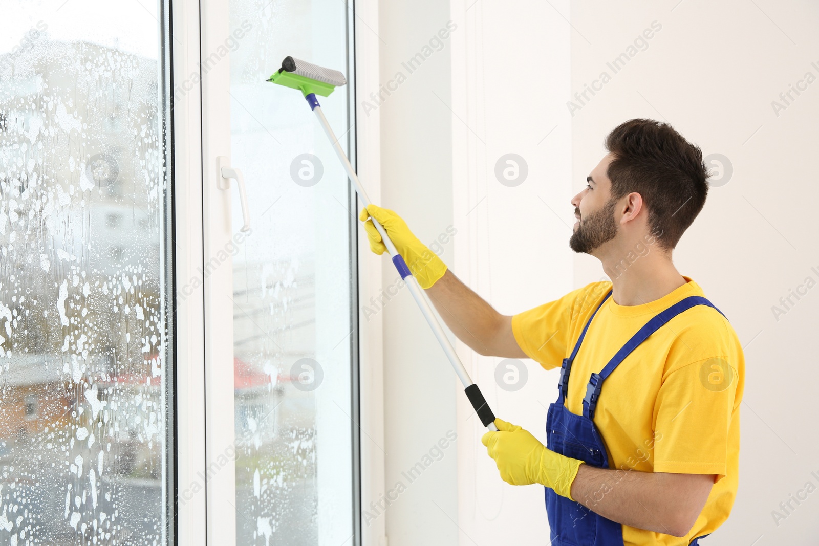 Photo of Professional young janitor in uniform cleaning window indoors