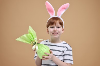 Photo of Easter celebration. Cute little boy with bunny ears and wrapped egg on dark beige background