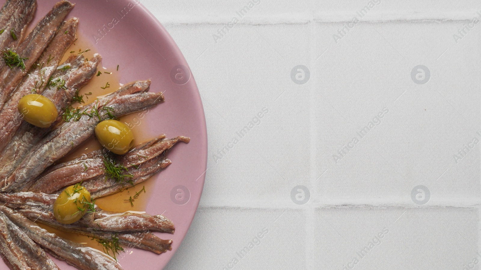 Photo of Canned anchovy fillets with olives on white tiled table, top view. Space for text