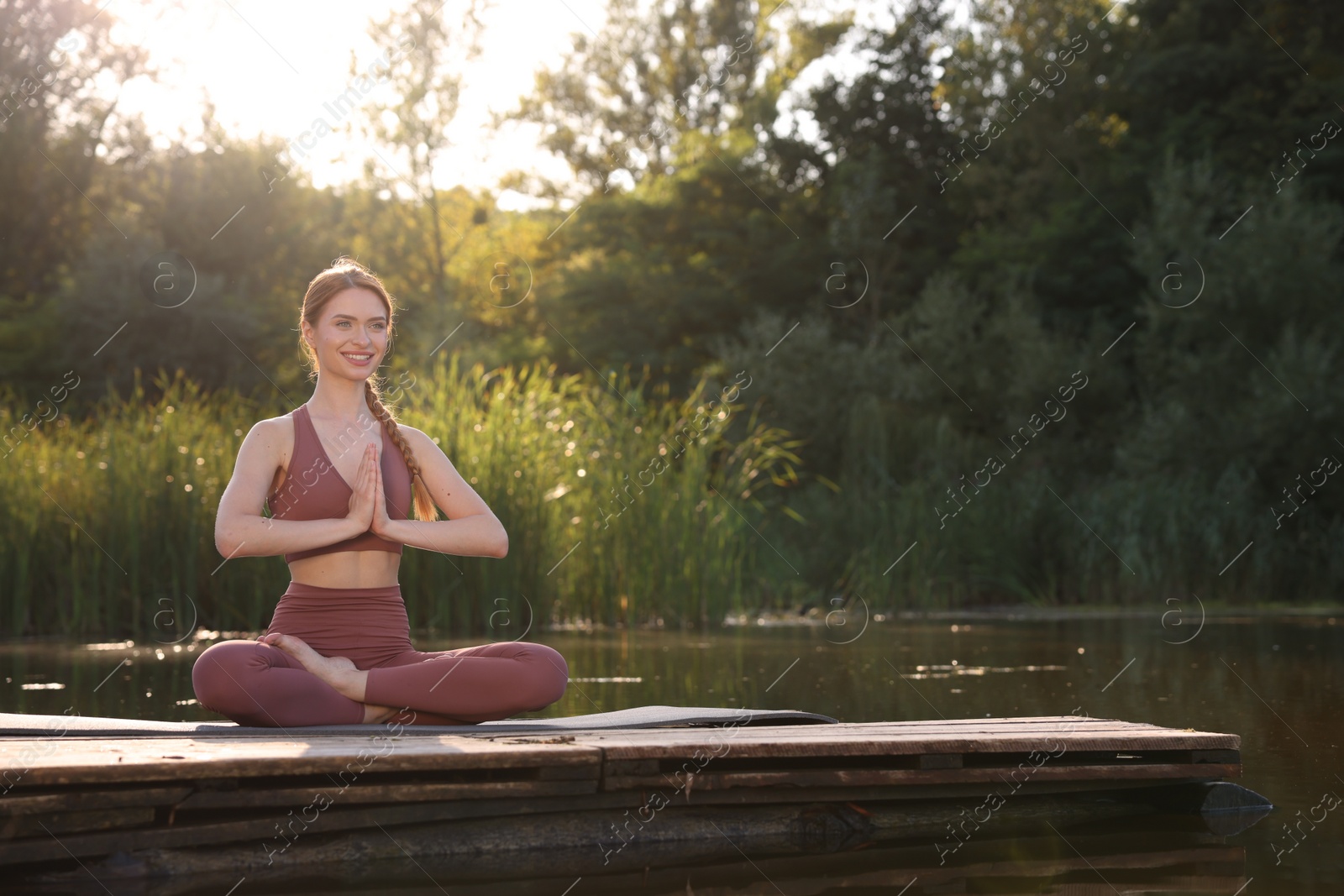 Photo of Beautiful happy woman practicing Padmasana on yoga mat on wooden pier near pond, space for text. Lotus pose