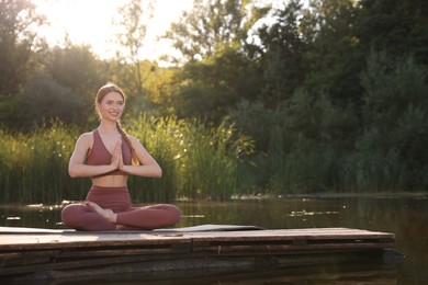 Beautiful happy woman practicing Padmasana on yoga mat on wooden pier near pond, space for text. Lotus pose