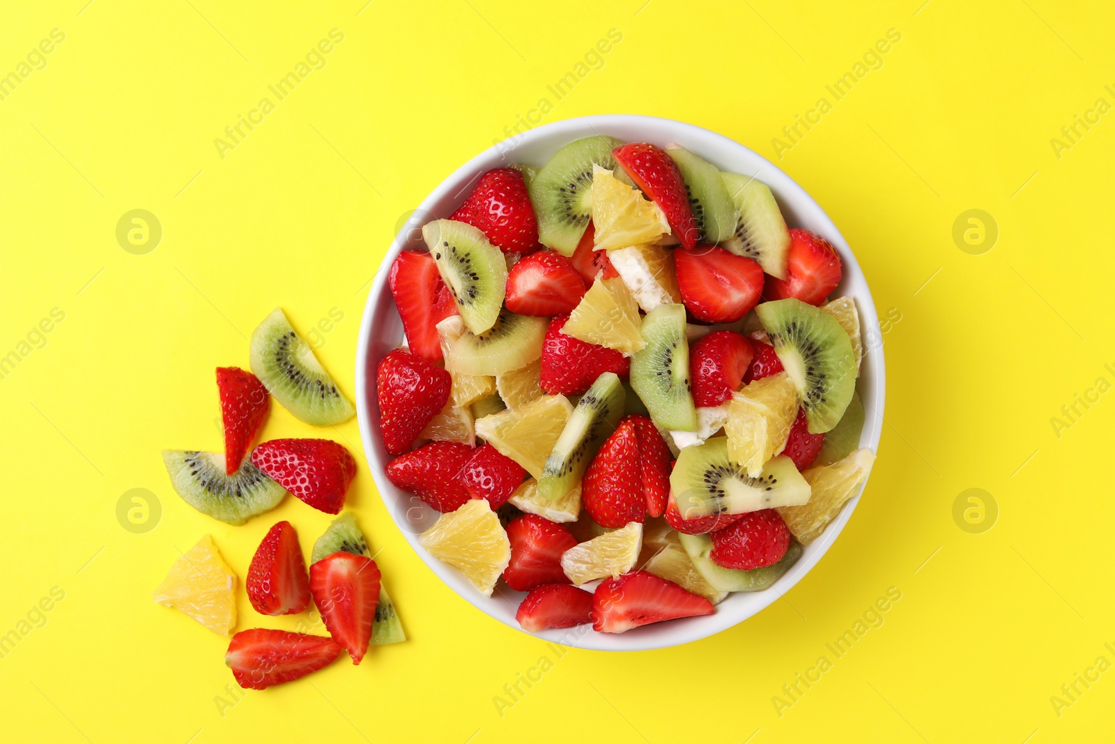 Photo of Yummy fruit salad in bowl and ingredients on yellow background, flat lay