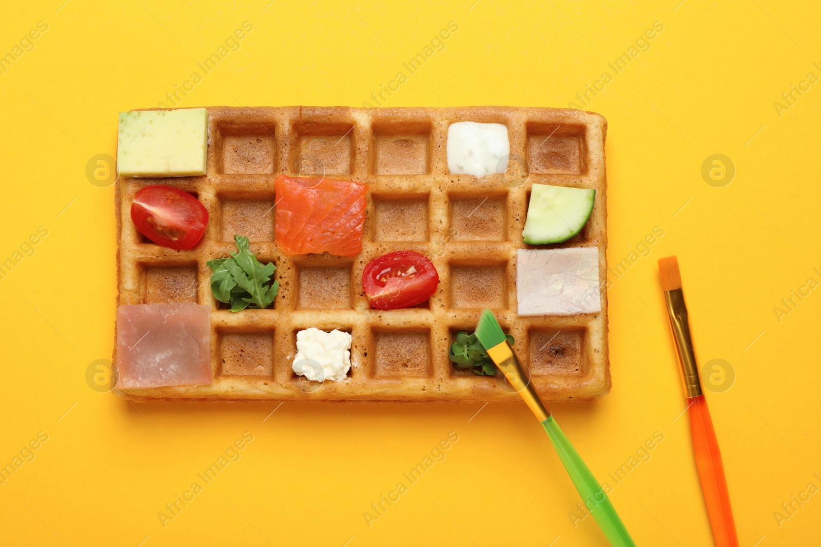 Photo of Brushes and waffle with different ingredients as paints on orange background, flat lay