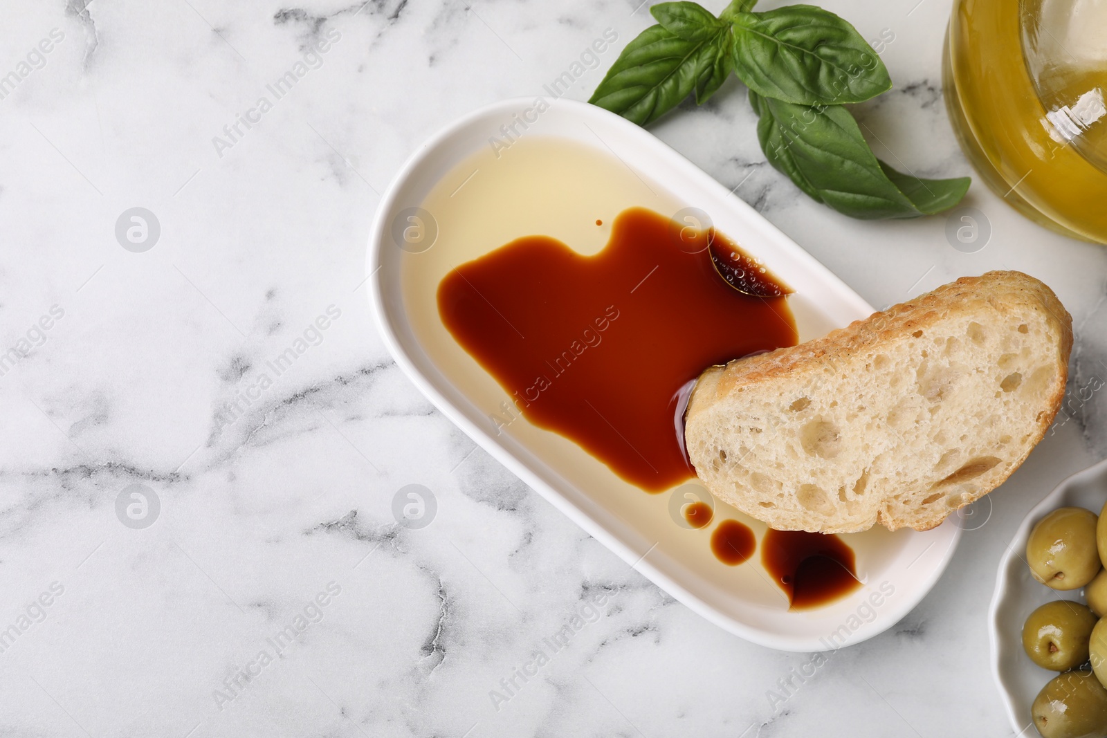 Photo of Bowl of organic balsamic vinegar with oil served with bread slice, basil and olives on white marble table, flat lay. Space for text