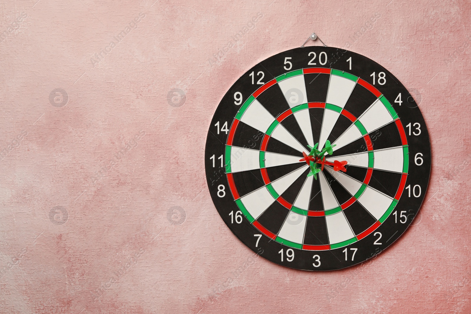 Photo of Dart board with arrows hitting target on pink wall. Space for text