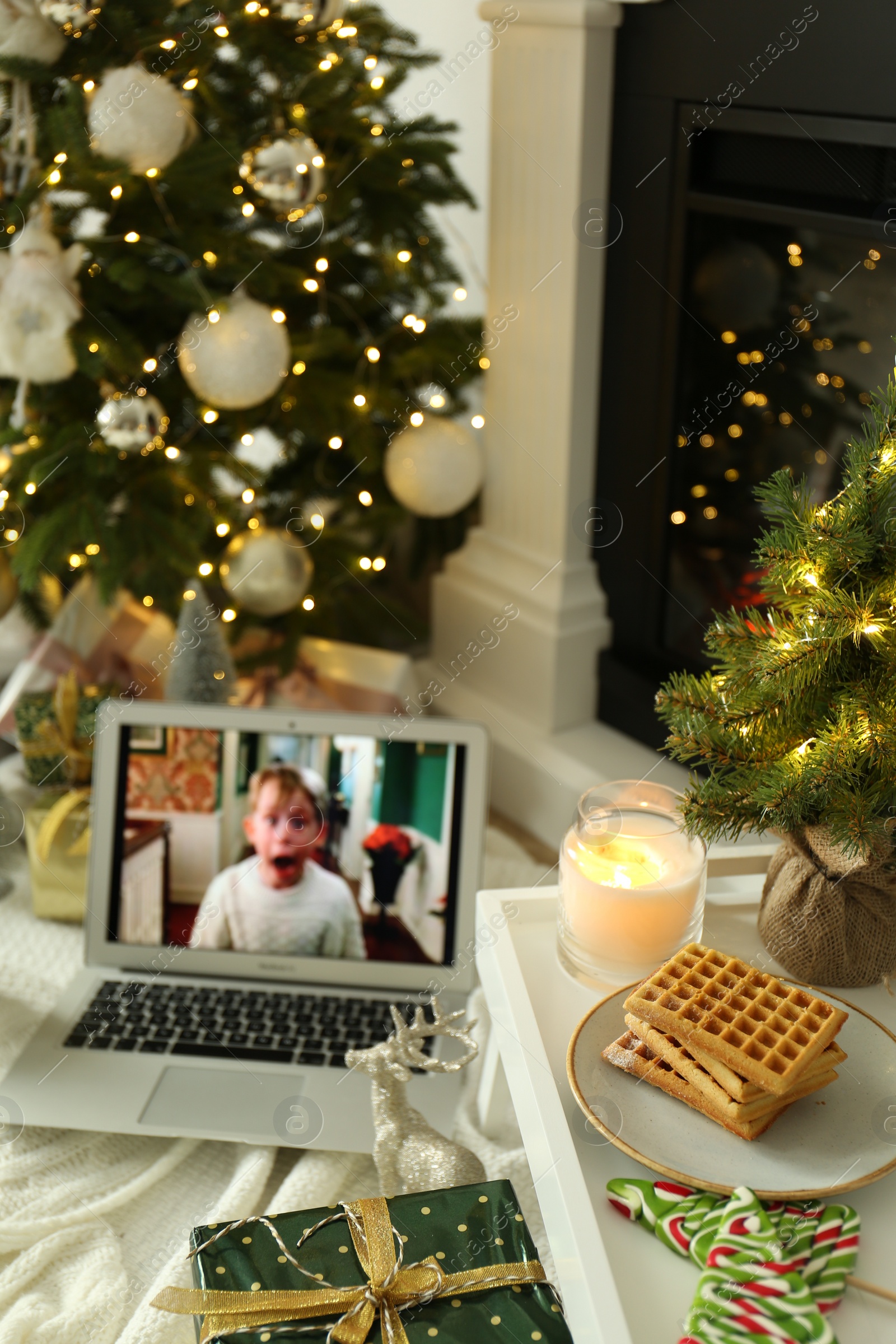 Photo of MYKOLAIV, UKRAINE - DECEMBER 23, 2020: Laptop displaying Home Alone movie near fireplace indoors, focus on tray with cookies and decor. Cozy winter holidays atmosphere