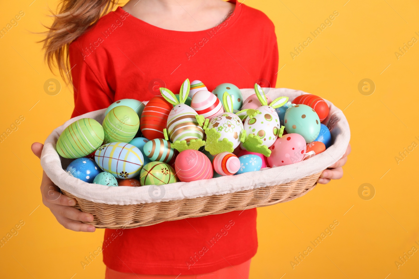 Photo of Little girl holding wicker basket full of Easter eggs on orange background, closeup