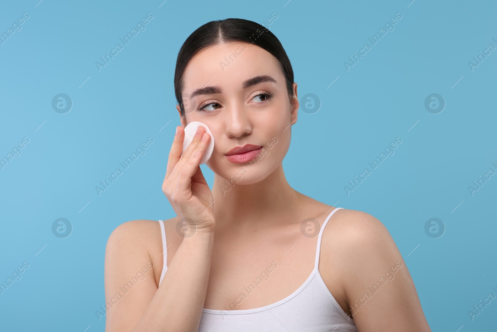 Photo of Beautiful woman removing makeup with cotton pad on light blue background