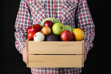 Photo of Man holding wooden crate filled with fresh vegetables and fruits against black background, closeup