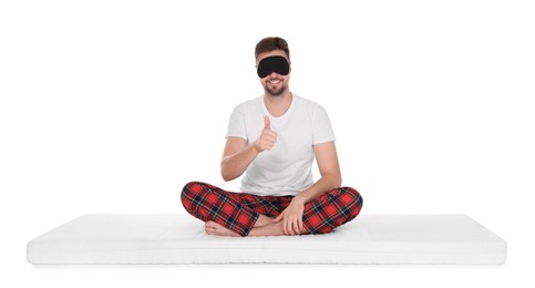 Photo of Man in sleeping mask sitting on soft mattress and showing thumb up against white background