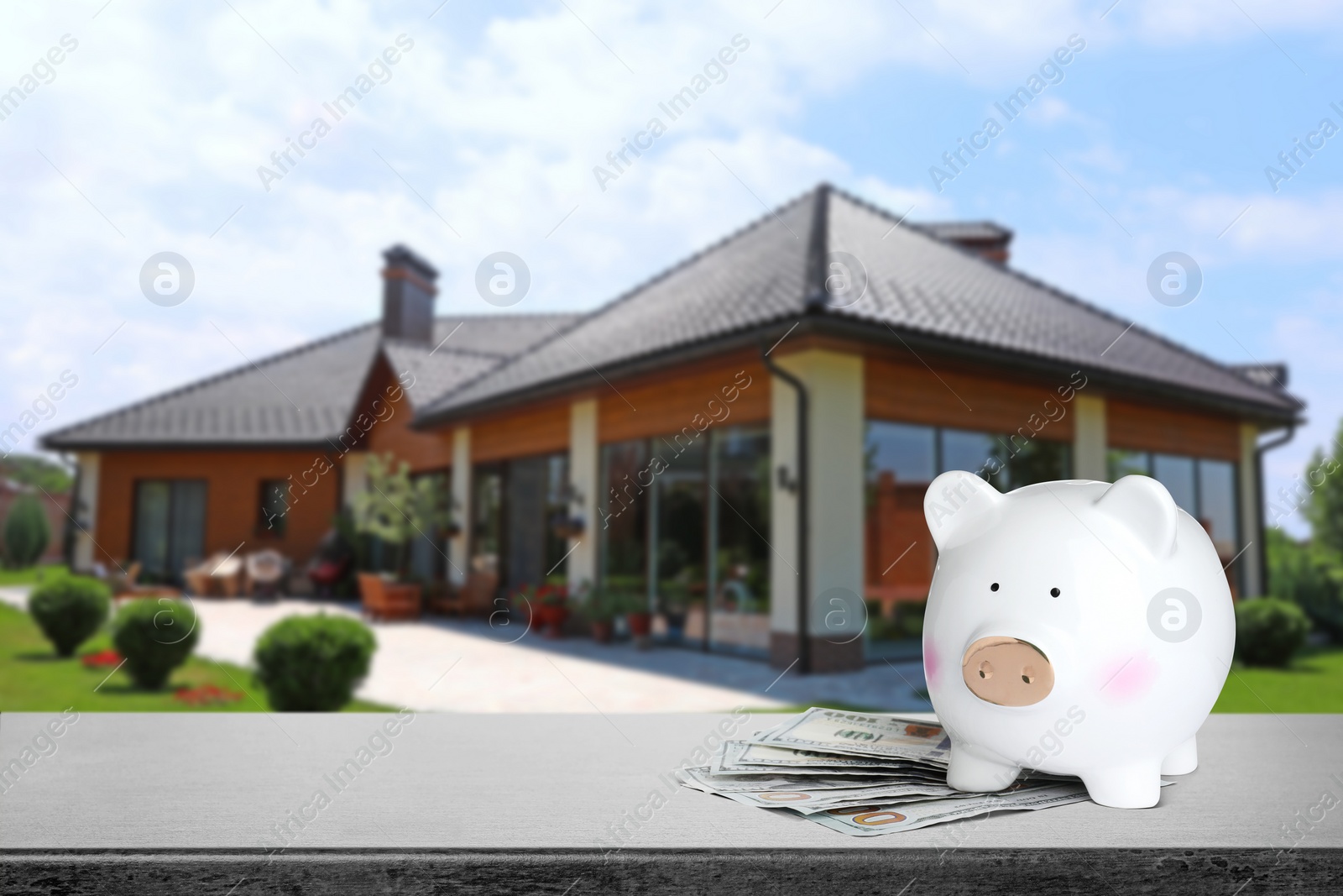 Image of Piggy bank and money on stone surface and blurred view of beautiful house, space for text. Mortgage concept