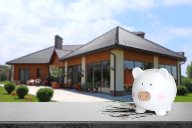 Image of Piggy bank and money on stone surface and blurred view of beautiful house, space for text. Mortgage concept