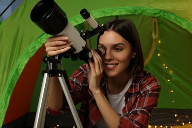 Photo of Young woman looking at stars through telescope while sitting in camping tent indoors