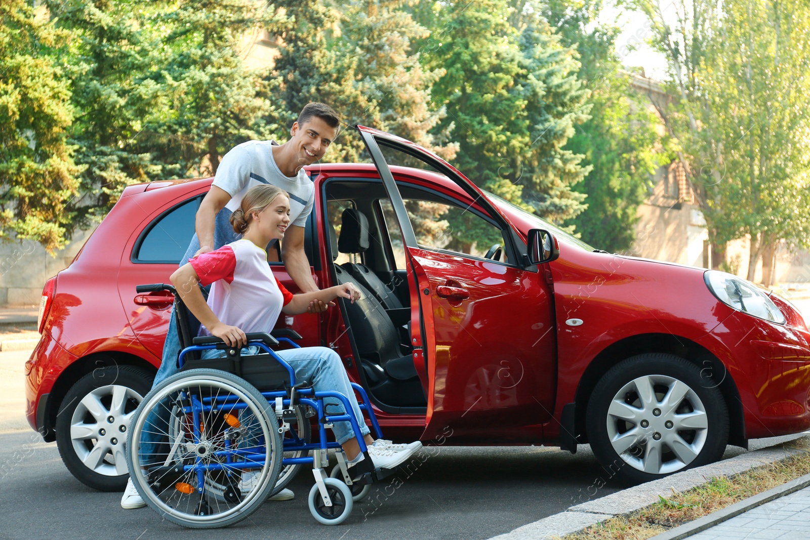 Photo of Young man helping disabled woman in wheelchair to get into car outdoors