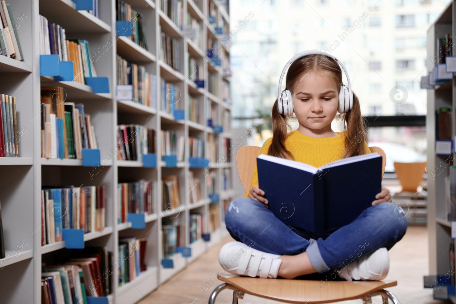 Photo of Cute little girl with headphones reading book on chair in library