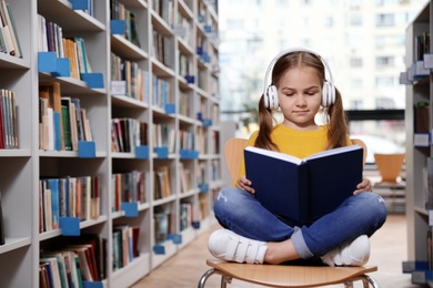 Cute little girl with headphones reading book on chair in library