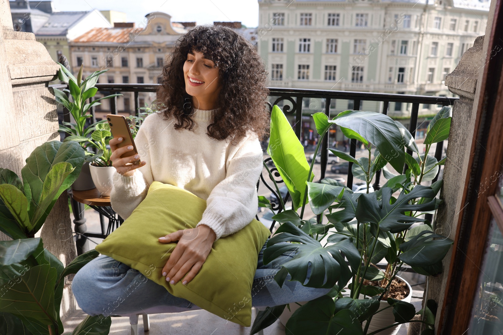 Photo of Beautiful young woman using smartphone surrounded by houseplants on balcony