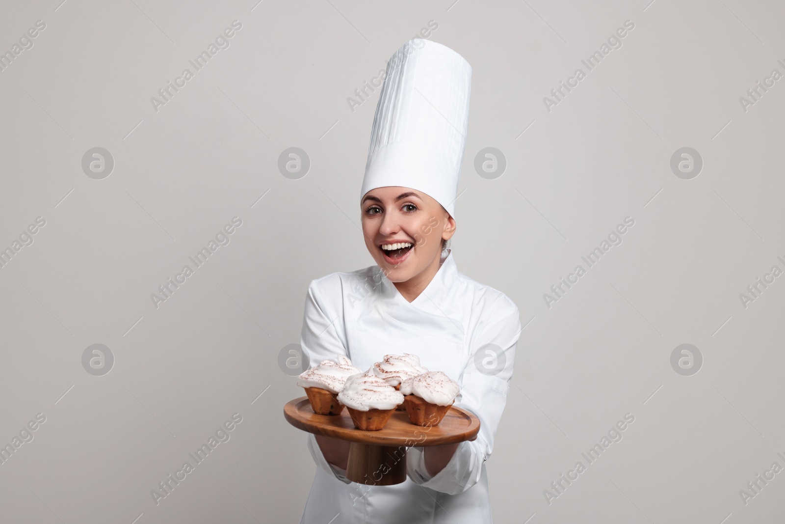 Photo of Happy professional confectioner in uniform holding delicious cupcakes on light grey background