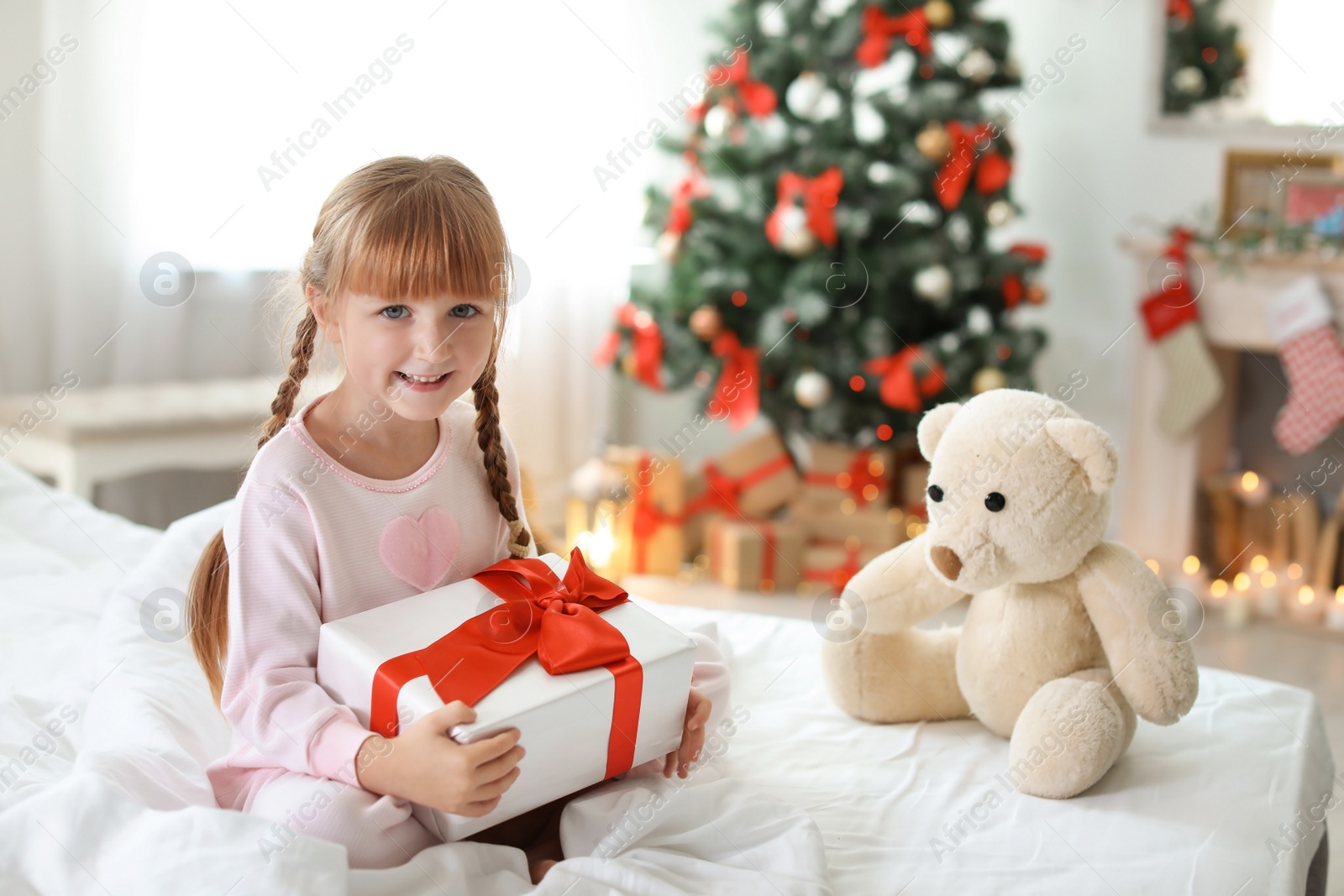 Photo of Cute little child with Christmas gift box sitting on bed at home