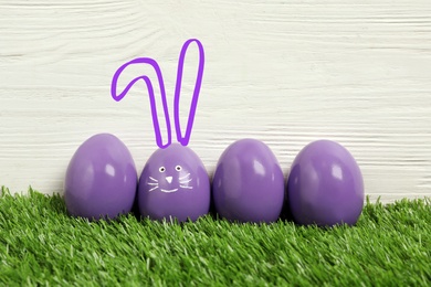 One egg with drawn face and ears as Easter bunny among others on green grass against white wooden background