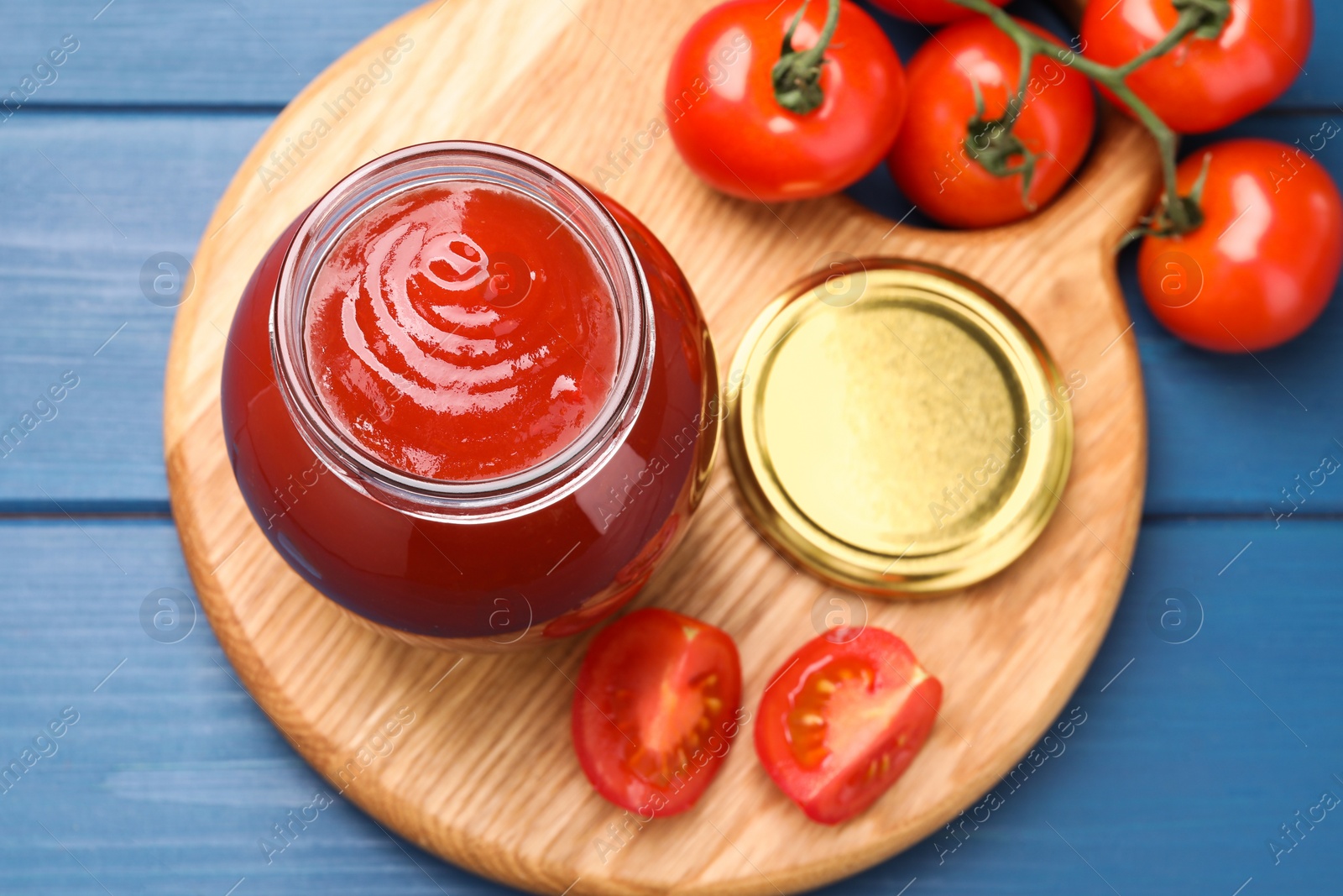 Photo of Organic ketchup in jar and fresh tomatoes on blue table, top view. Tomato sauce