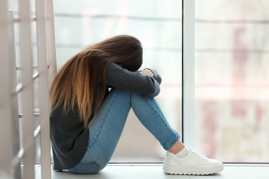 Photo of Upset teenage girl sitting at window indoors. Space for text