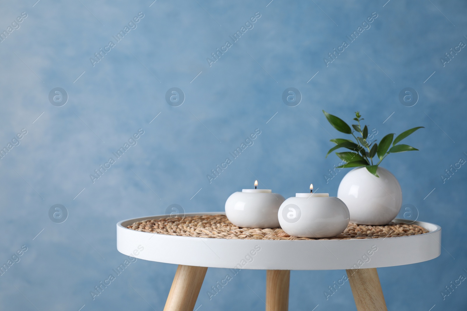 Photo of Burning candles and plant on white table against light blue background, space for text