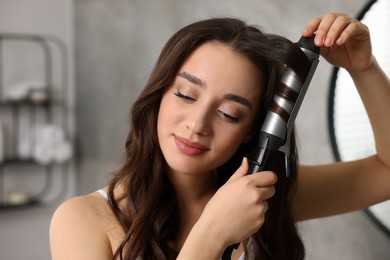 Photo of Beautiful woman using curling hair iron in bathroom