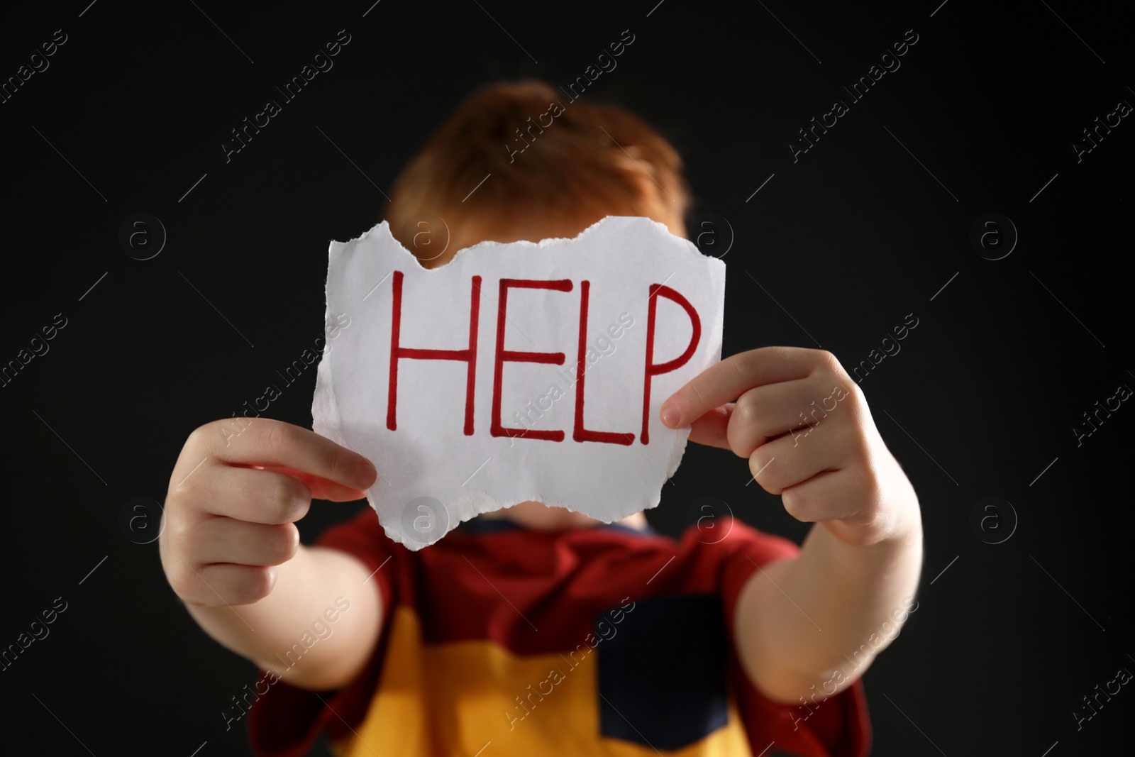 Photo of Little boy holding piece of paper with word Help against black background, focus on hands. Domestic violence concept