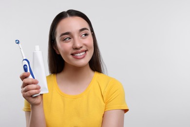 Happy young woman holding electric toothbrush and tube of toothpaste on white background