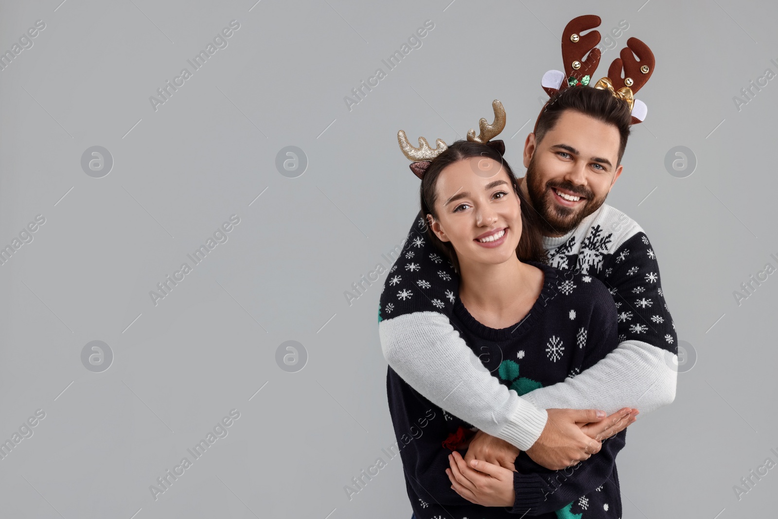 Photo of Happy young couple in Christmas sweaters and reindeer headbands on grey background. Space for text