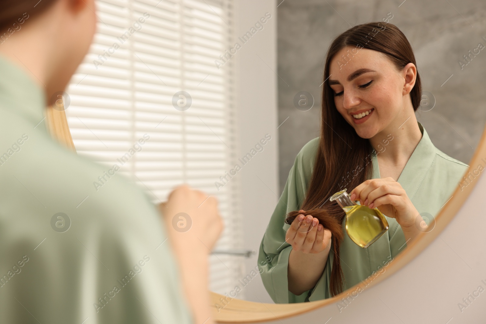 Photo of Young woman applying oil hair mask near mirror at home