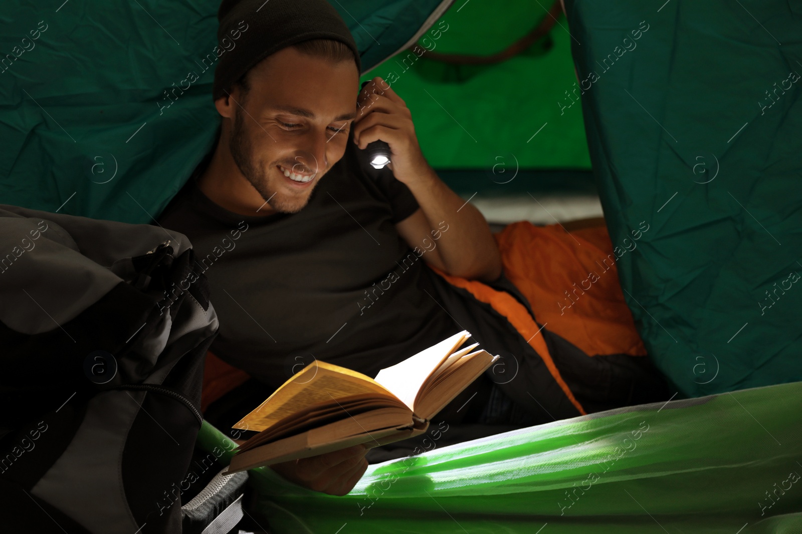 Photo of Young man in sleeping bag reading book with flashlight inside of tent