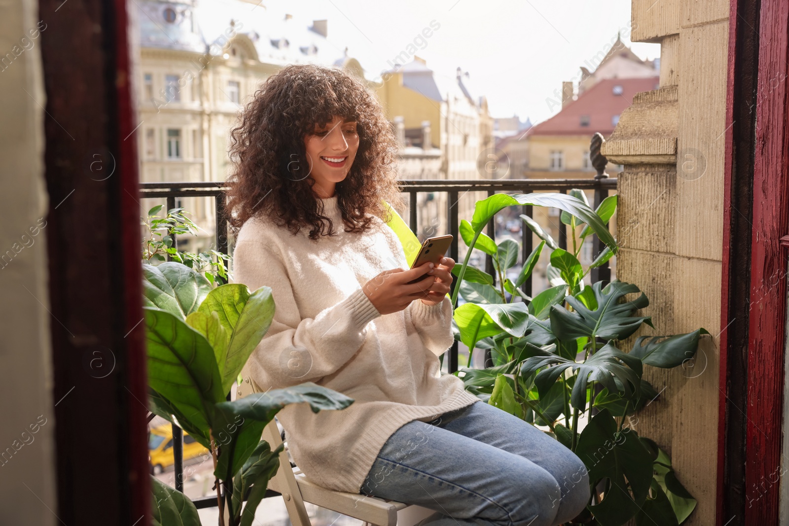 Photo of Beautiful young woman using smartphone surrounded by houseplants on balcony