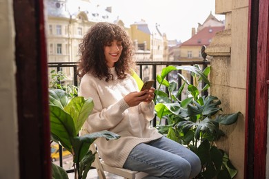 Beautiful young woman using smartphone surrounded by houseplants on balcony