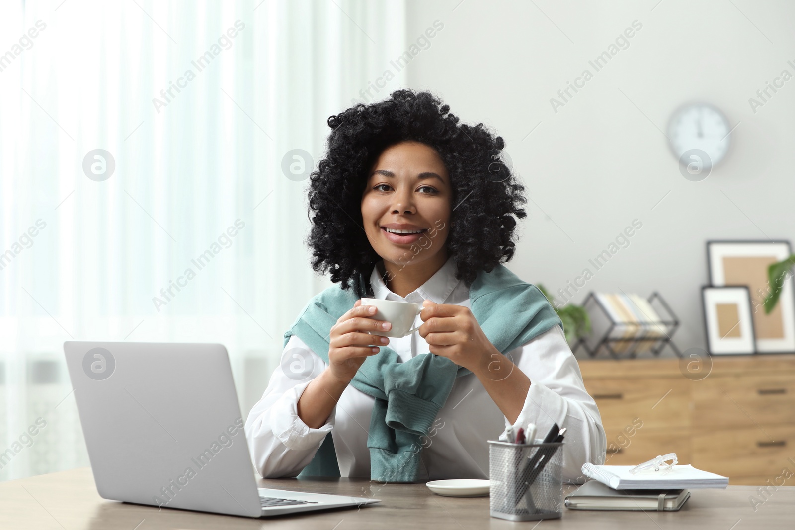 Photo of Happy young woman with cup of drink using laptop at wooden desk indoors