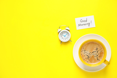 Photo of Delicious coffee, alarm clock and card with GOOD MORNING wish on yellow background, flat lay. Space for text