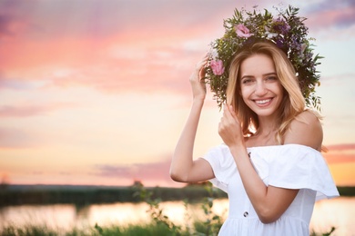 Photo of Young woman wearing wreath made of beautiful flowers outdoors at sunset