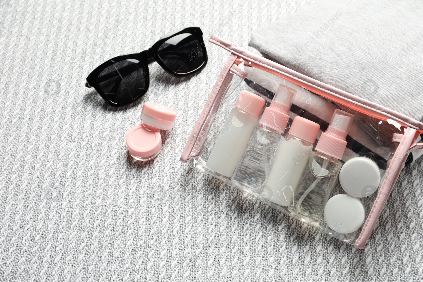 Photo of Cosmetic travel kit. Plastic bag with small containers of personal care products, stack of clothes and sunglasses on sofa