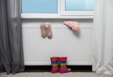 Photo of Heating radiator with knitted hat, socks and rubber boots near window indoors