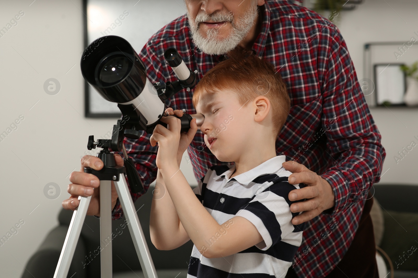 Photo of Little boy with his grandfather looking at stars through telescope in room