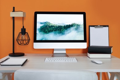 Photo of Modern computer, books, lamp and notebook on wooden desk near orange wall. Home office